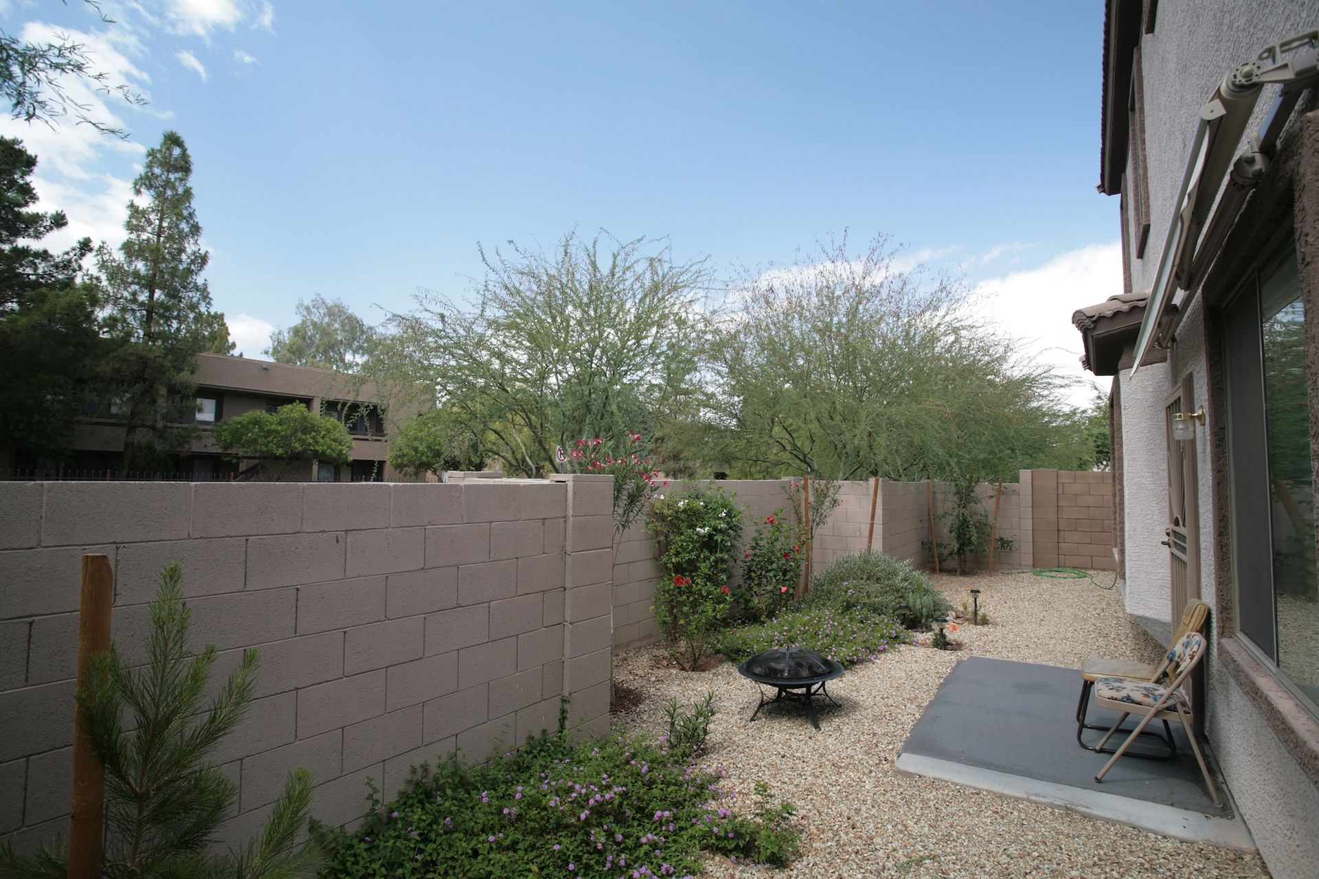 Backyard with gravel ground, concrete block walls, greenery, and a fire pit under a blue sky.
