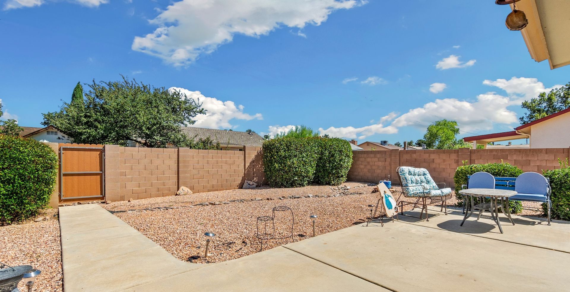 Backyard patio with chairs, gravel, and a brown wall under a blue sky.