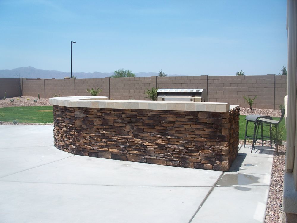 Outdoor stone-covered bar with countertop on a concrete patio, grill in the background, blue sky.