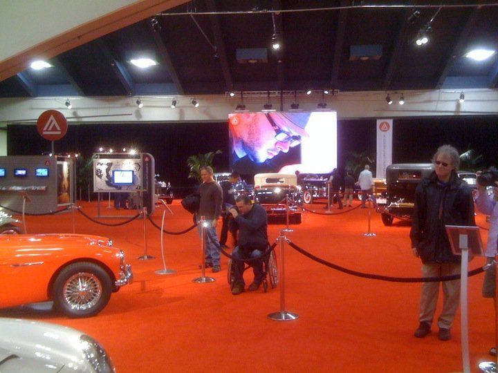 An orange car and other vintage vehicles on display at a car show with people viewing.