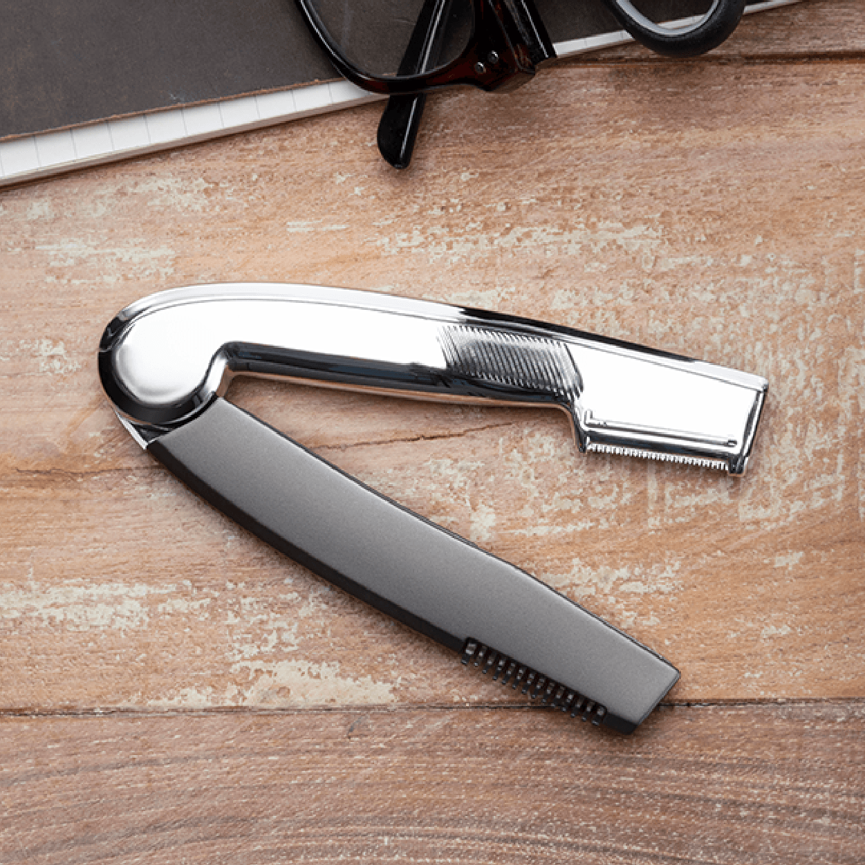 A silver and black straight razor on a wooden surface, near a pair of glasses and a book.