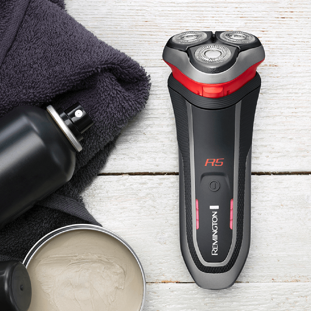 Black and red Remington F5 electric shaver with shaving cream, spray, and towel on a wood table.