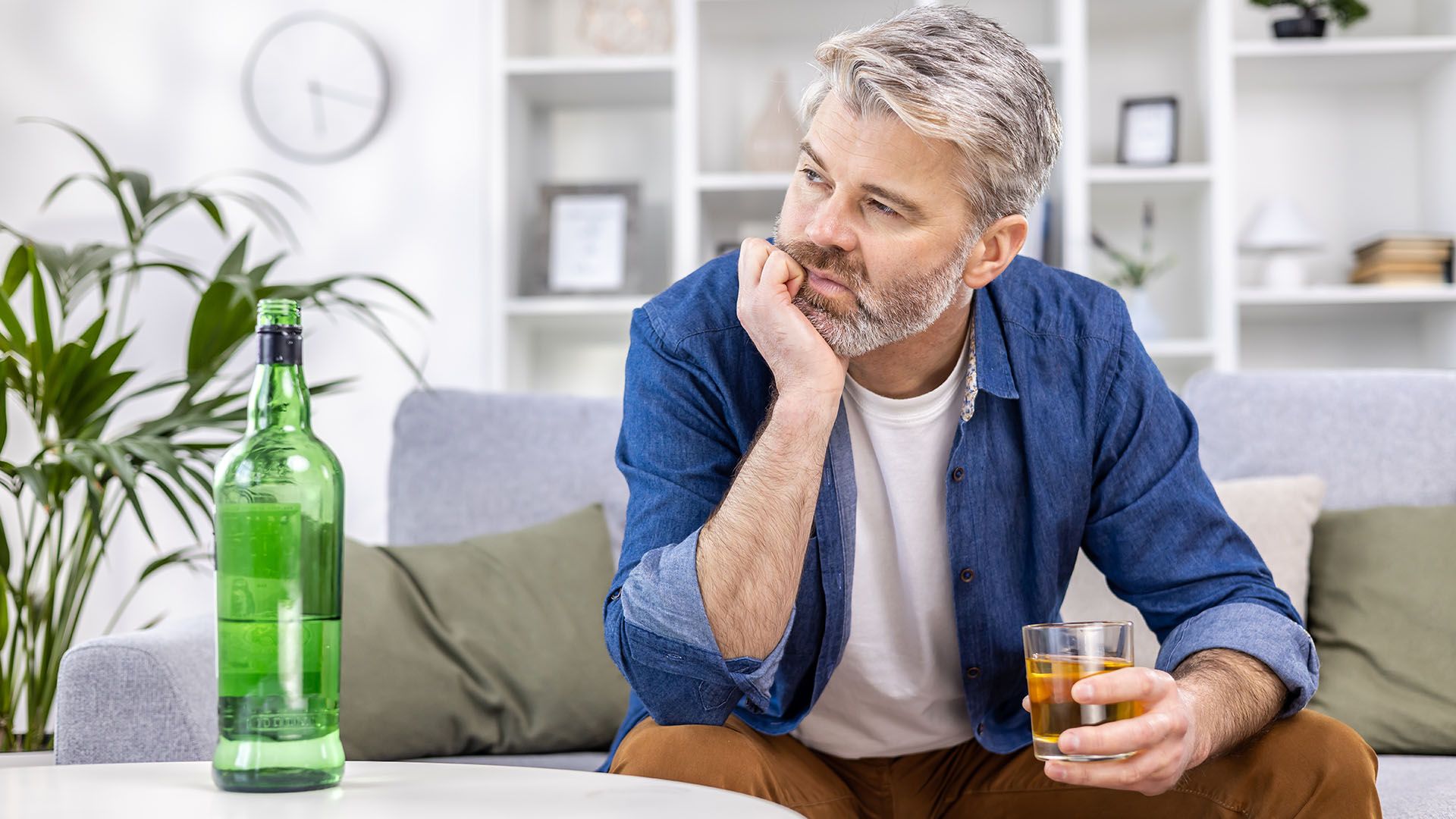 A man is sitting on a couch holding a glass and a bottle of alcohol.