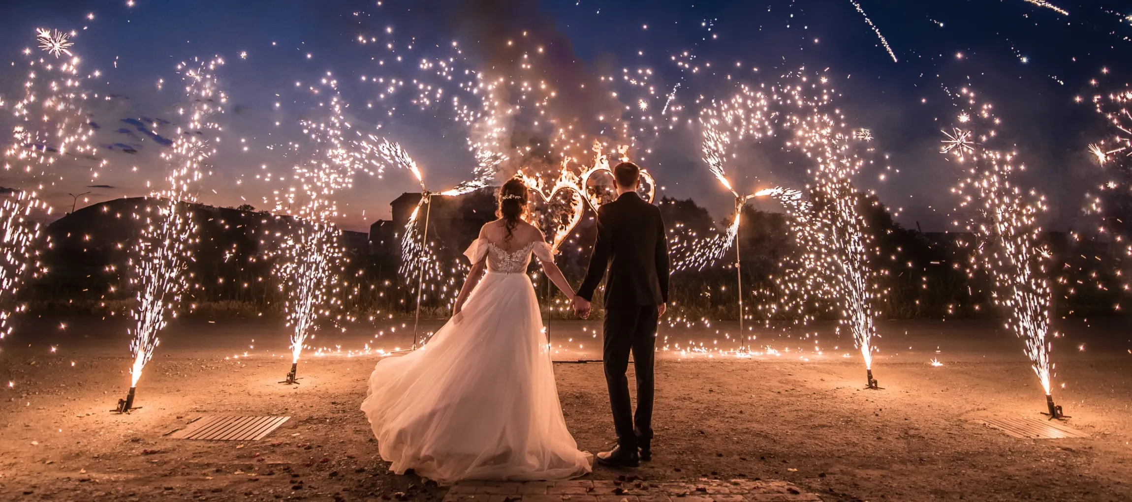 A bride and groom are holding hands in front of fireworks.