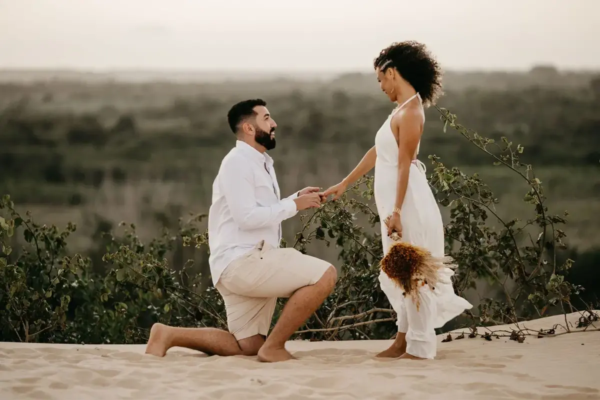 A man is kneeling down to propose to a woman on a sand dune.