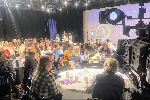 A large group of people are sitting at tables in front of a camera.