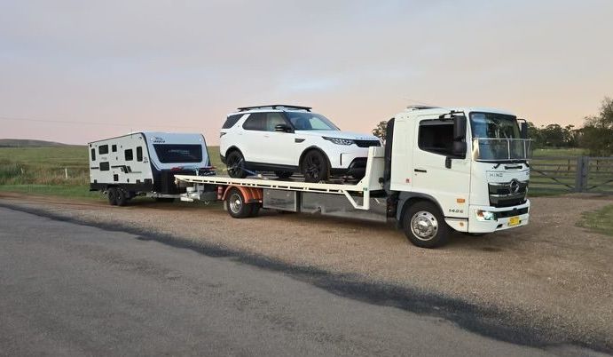 A Tow Truck Is Carrying A White Car On Its Flatbed and Towing A White Caravan — NCM Towing Newcastle In Broadmeadow, NSW