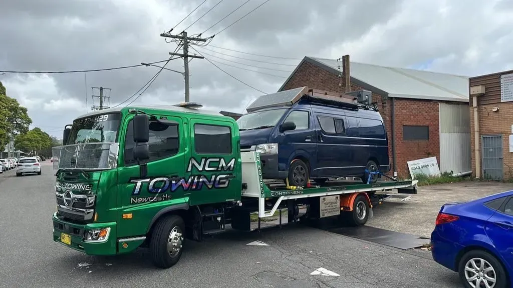 A Green Towing Truck Is Carrying A Blue Van Down A Street — NCM Towing Newcastle In Central Coast, NSW