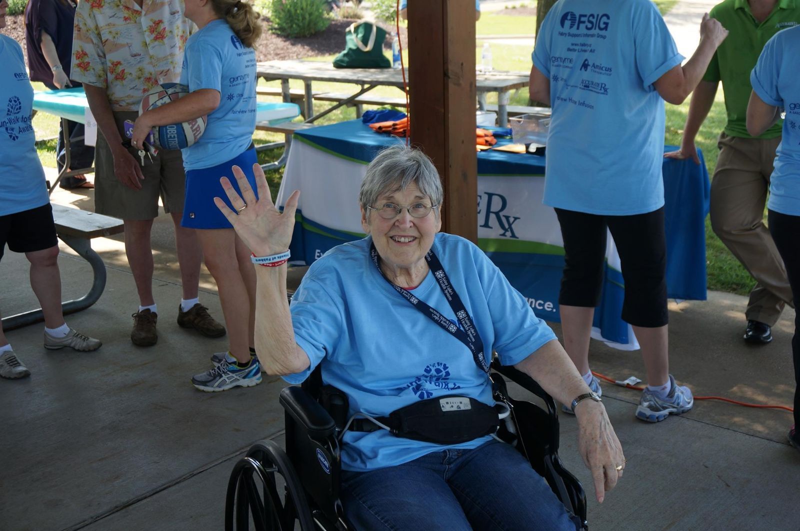 Woman in wheelchair waving at an outdoor event, wearing a blue shirt.