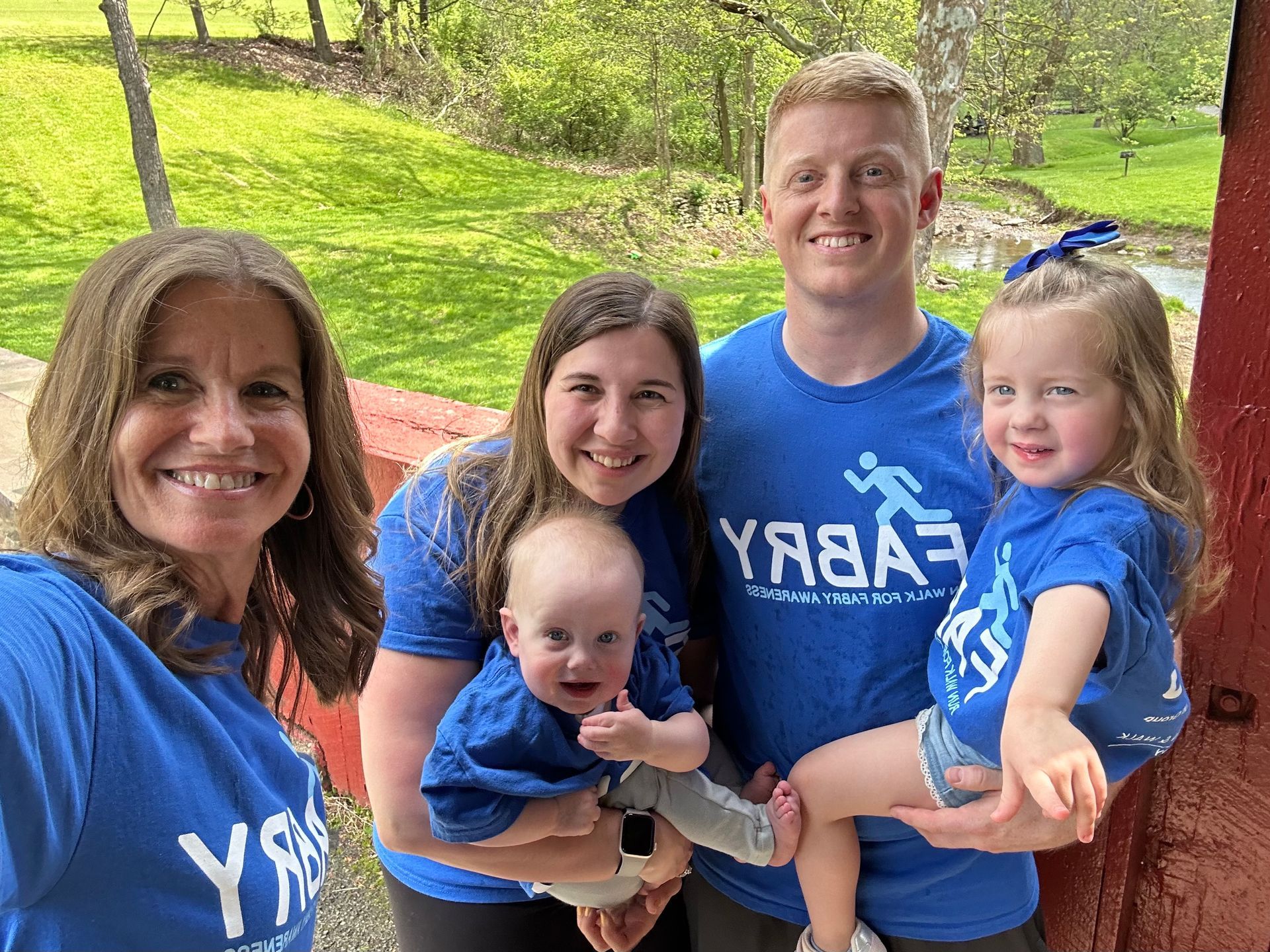 Family in blue shirts smiles outdoors. Baby held by woman, two other children. Fabry logo.