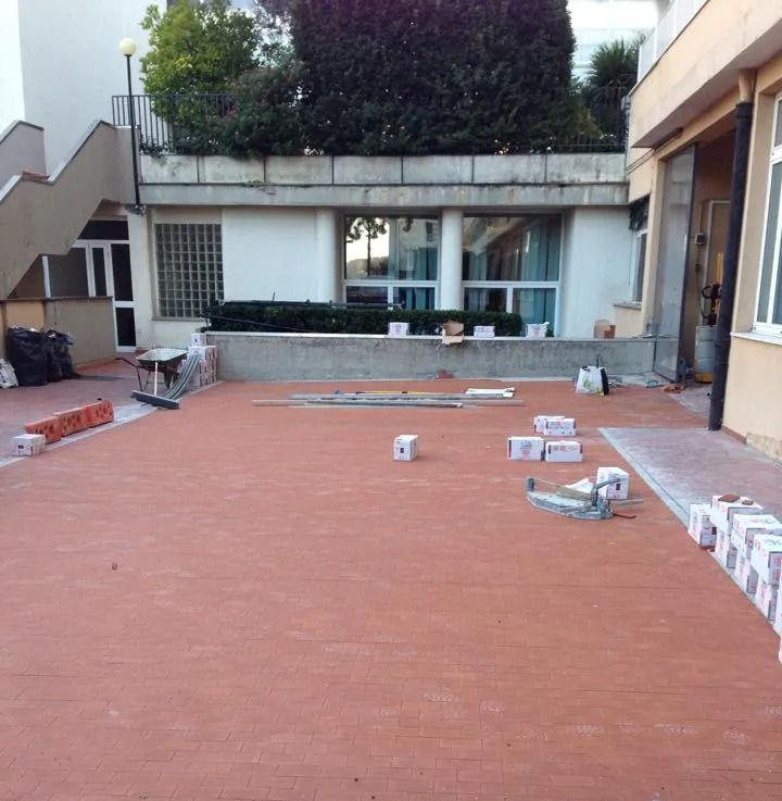Cortile esterno con pavimentazione in piastrelle rosse, materiali edili sparsi e la facciata di un edificio sullo sfondo.