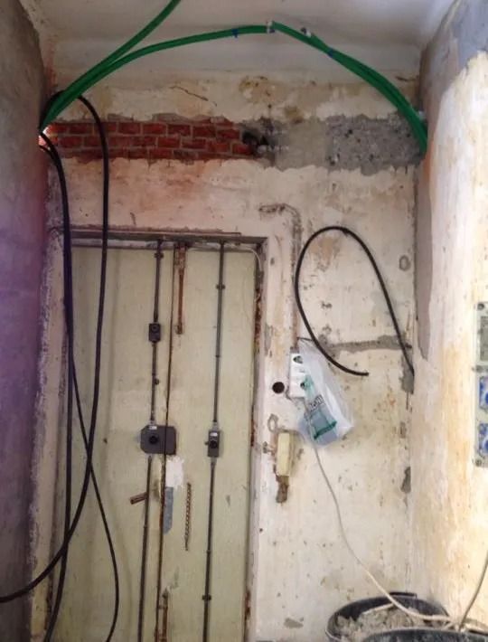 Parete danneggiata con tubi e cavi a vista e una piccola porta in uno stretto locale di servizio interno.