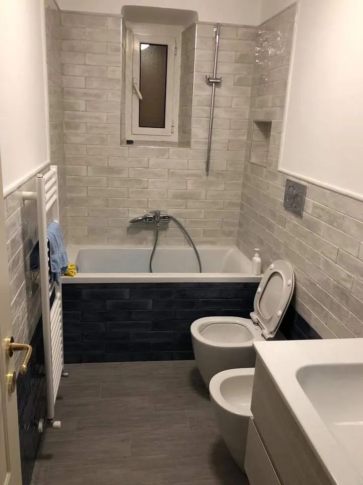Piccolo bagno piastrellato con vasca da bagno, WC, lavandino, termosifone e una piccola finestra sopra la vasca.