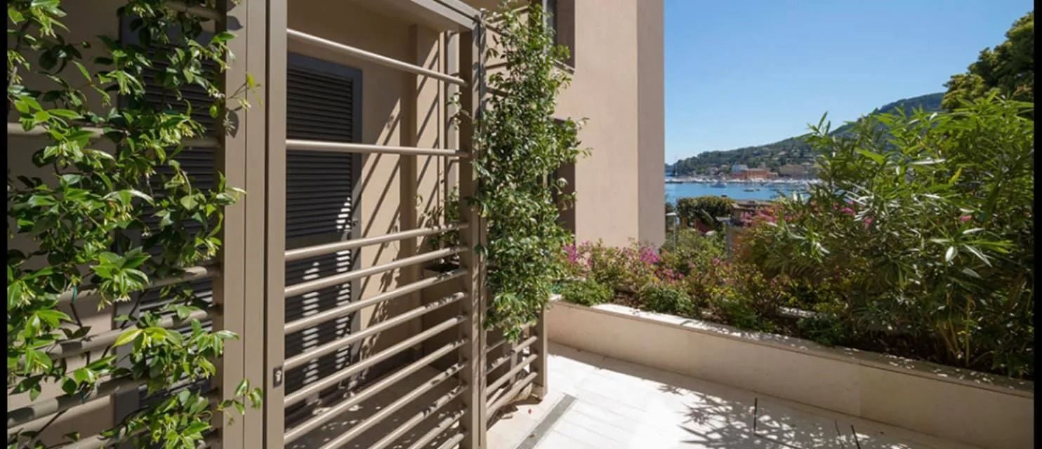 Balcone con persiane beige e piante rampicanti verdi, affacciato su una collina soleggiata e su una vista sull'acqua azzurra.