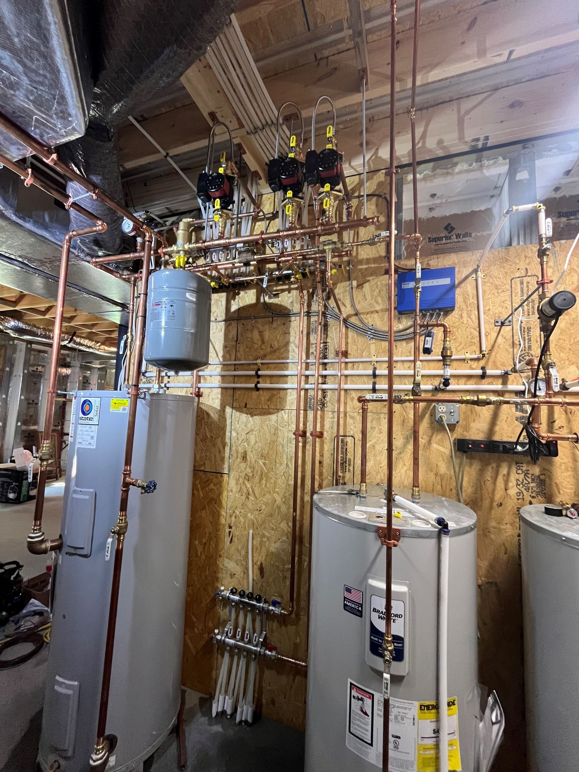 Plumbing installation in a utility room with copper pipes, three water heaters, and various valves against a wood-paneled wall.