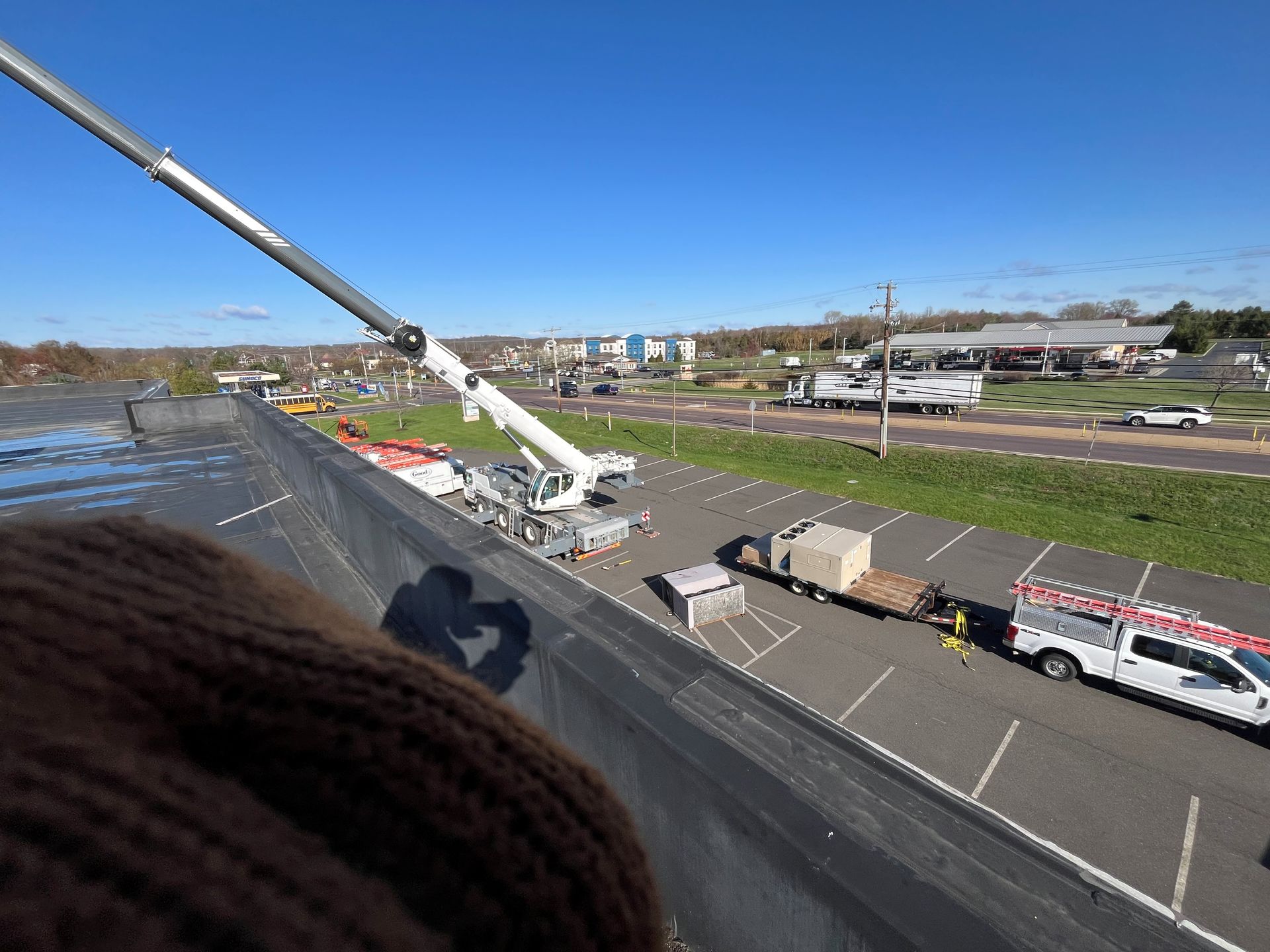 Crane lifting materials onto a flat roof on a sunny day. Parking lot and distant buildings visible.