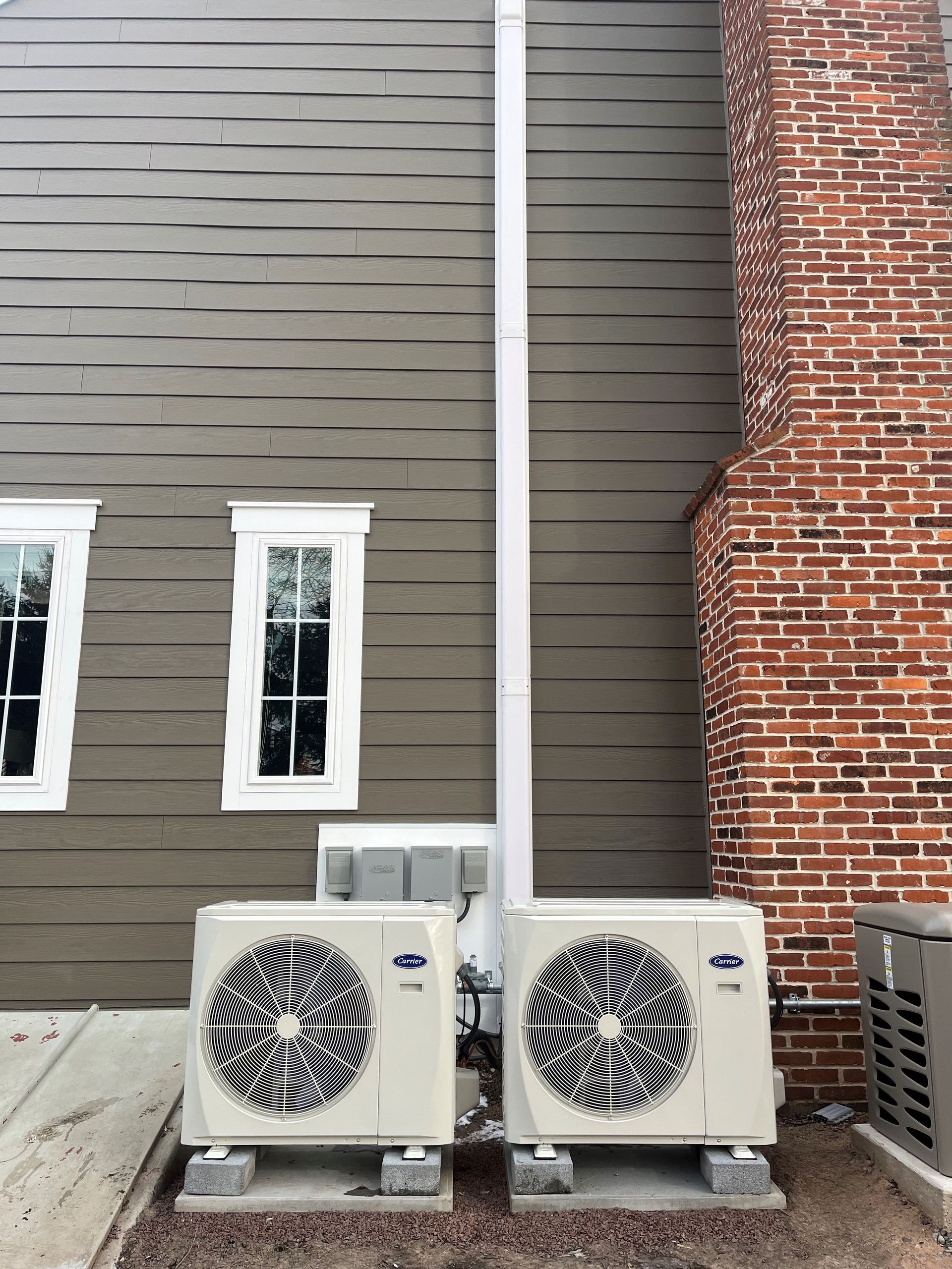 Two HVAC units next to a building with windows, chimney, and siding.