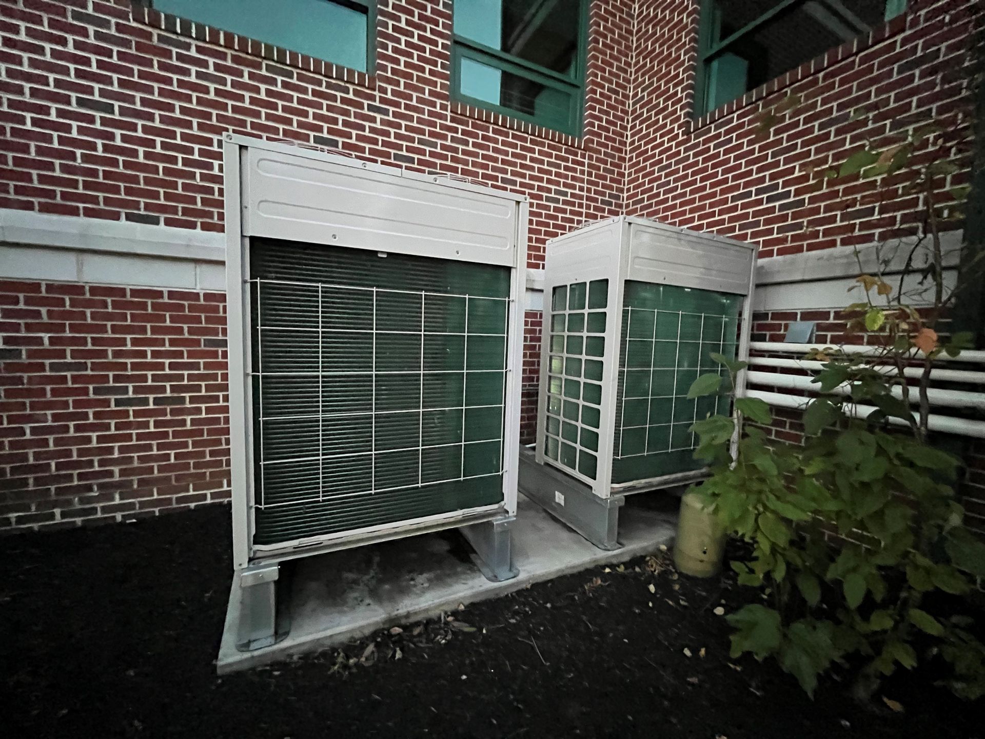 Two outdoor HVAC units near a brick building, with green metal grates and a small plant.