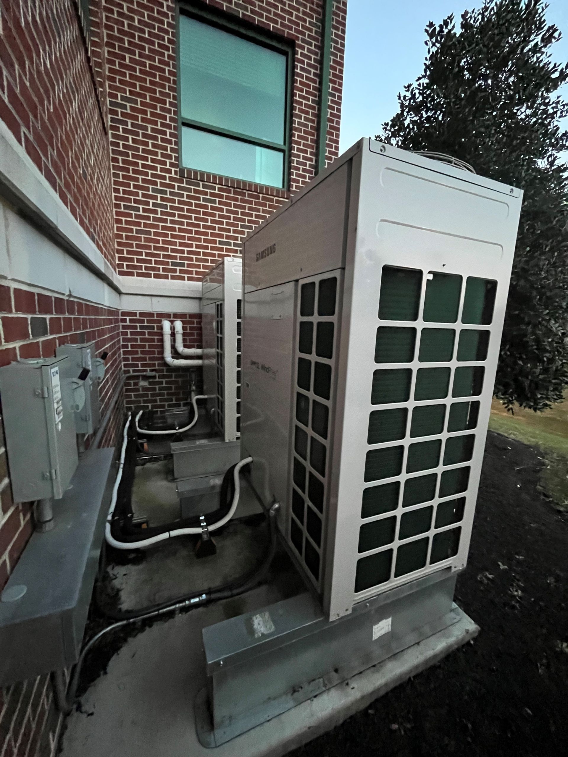HVAC unit outside a brick building with a window. Electrical boxes and pipes are visible.