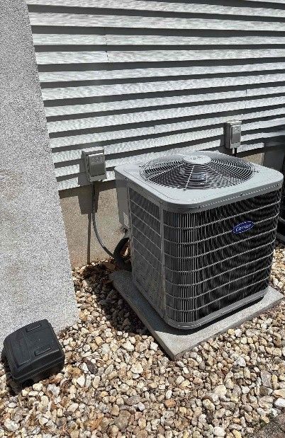 An air conditioner unit on a concrete pad next to a house with grey siding. Pebbles are on the ground.