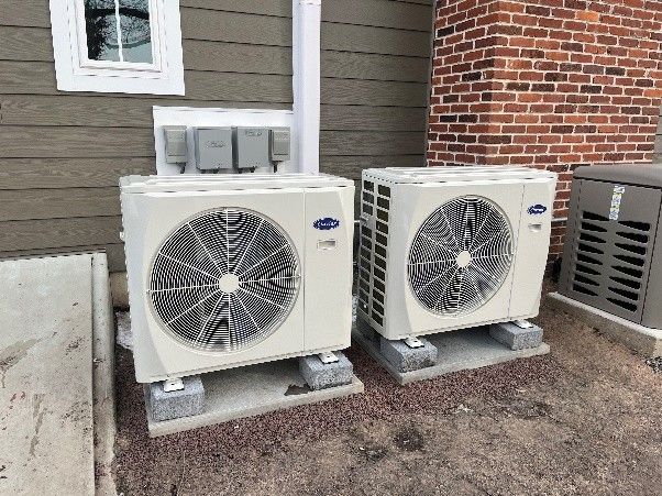 Two white Carrier air conditioning units on concrete blocks near a brick wall.