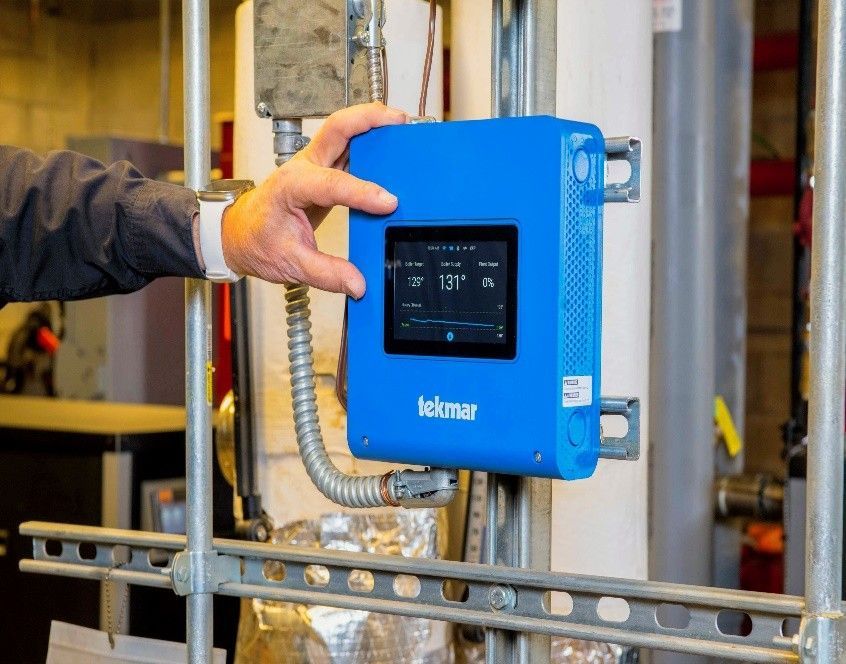 A person holding a blue Tekmar control panel on a metal pipe in an industrial setting.