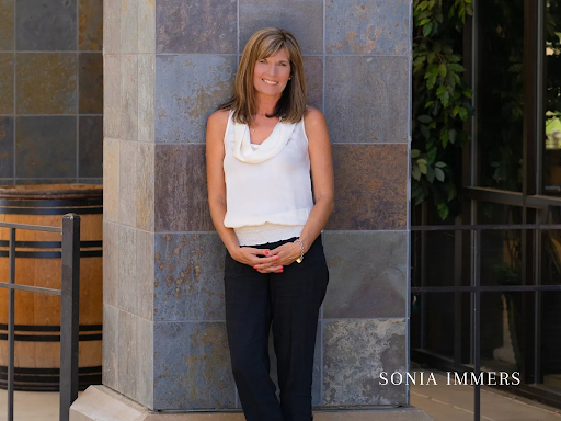 A woman in a white tank top and black pants is leaning against a stone wall.