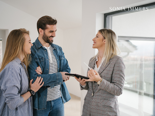 A realtor shows a smiling couple a property. The woman holds a tablet, the setting is modern.