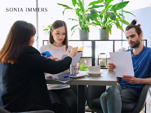 Three people reviewing documents at a table. Plants in the background, likely a cafe.