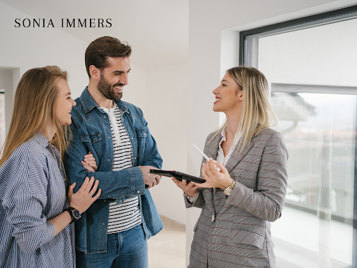 Real estate agent with couple looking at tablet, smiling in a light-filled room.