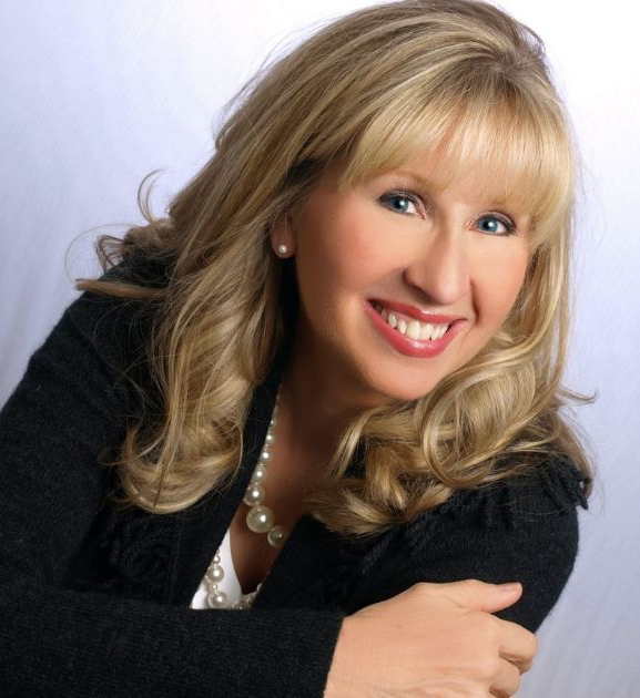A smiling woman with shoulder-length blonde hair, wearing a black textured jacket and a necklace, posed against white.