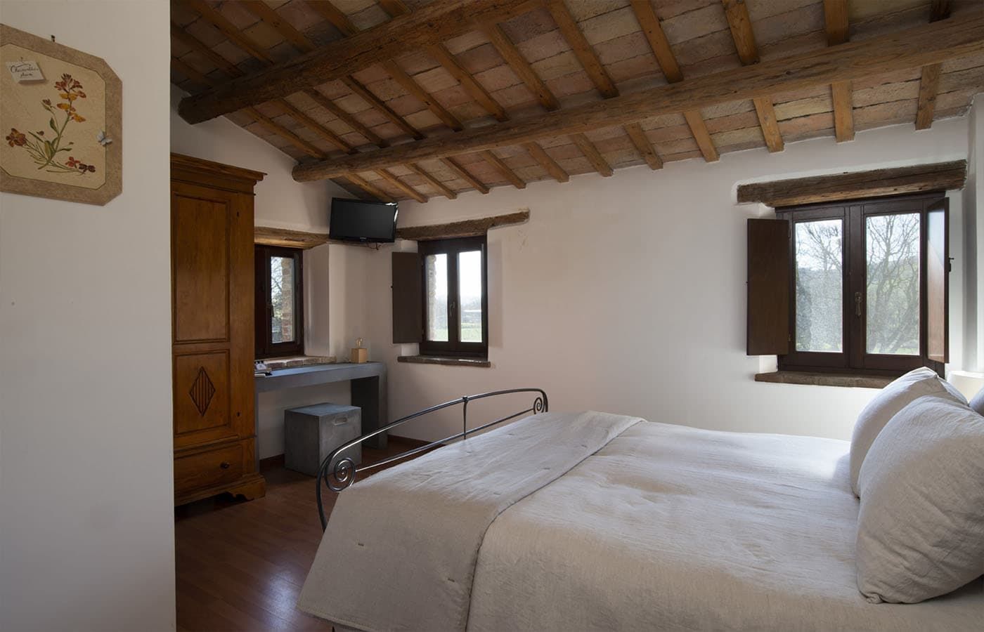 Camera da letto rustica con letto bianco, travi in legno a vista e due finestre che si affacciano sugli alberi.