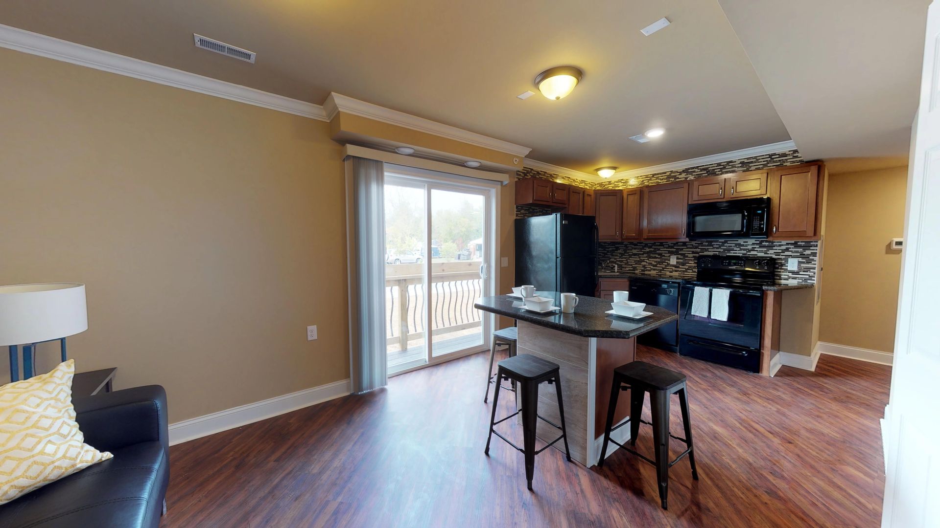 A living room with a couch , table and stools and a kitchen with a sliding glass door.