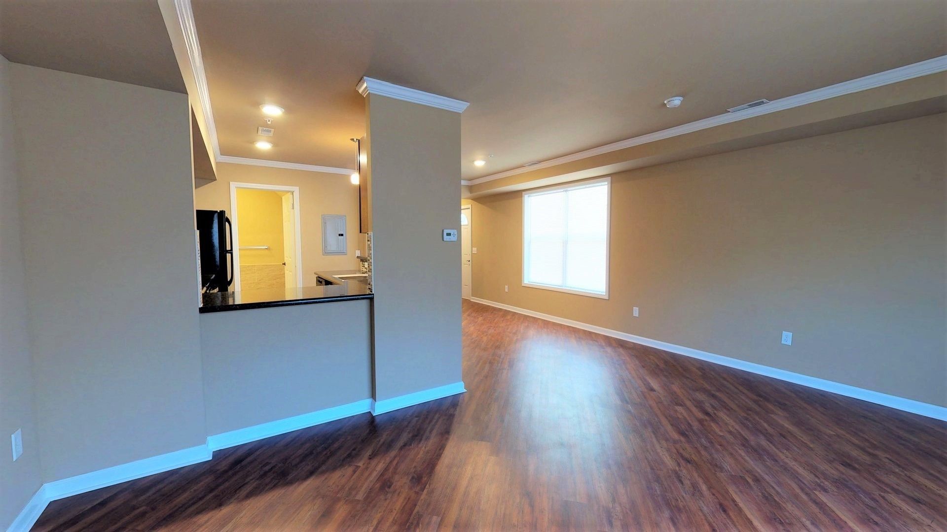 An empty living room with hardwood floors and a kitchen in the background.