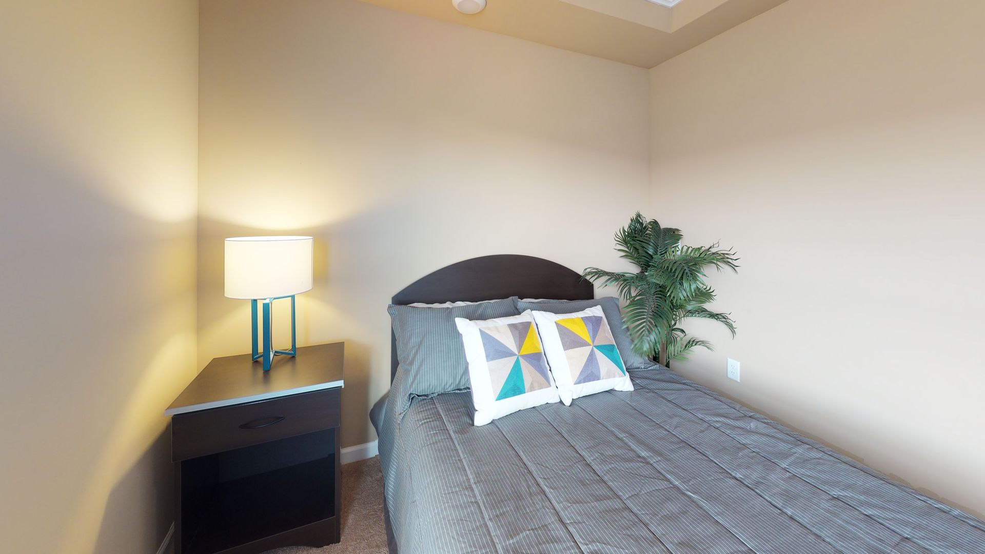 A bedroom with a bed , nightstand , lamp and plant.