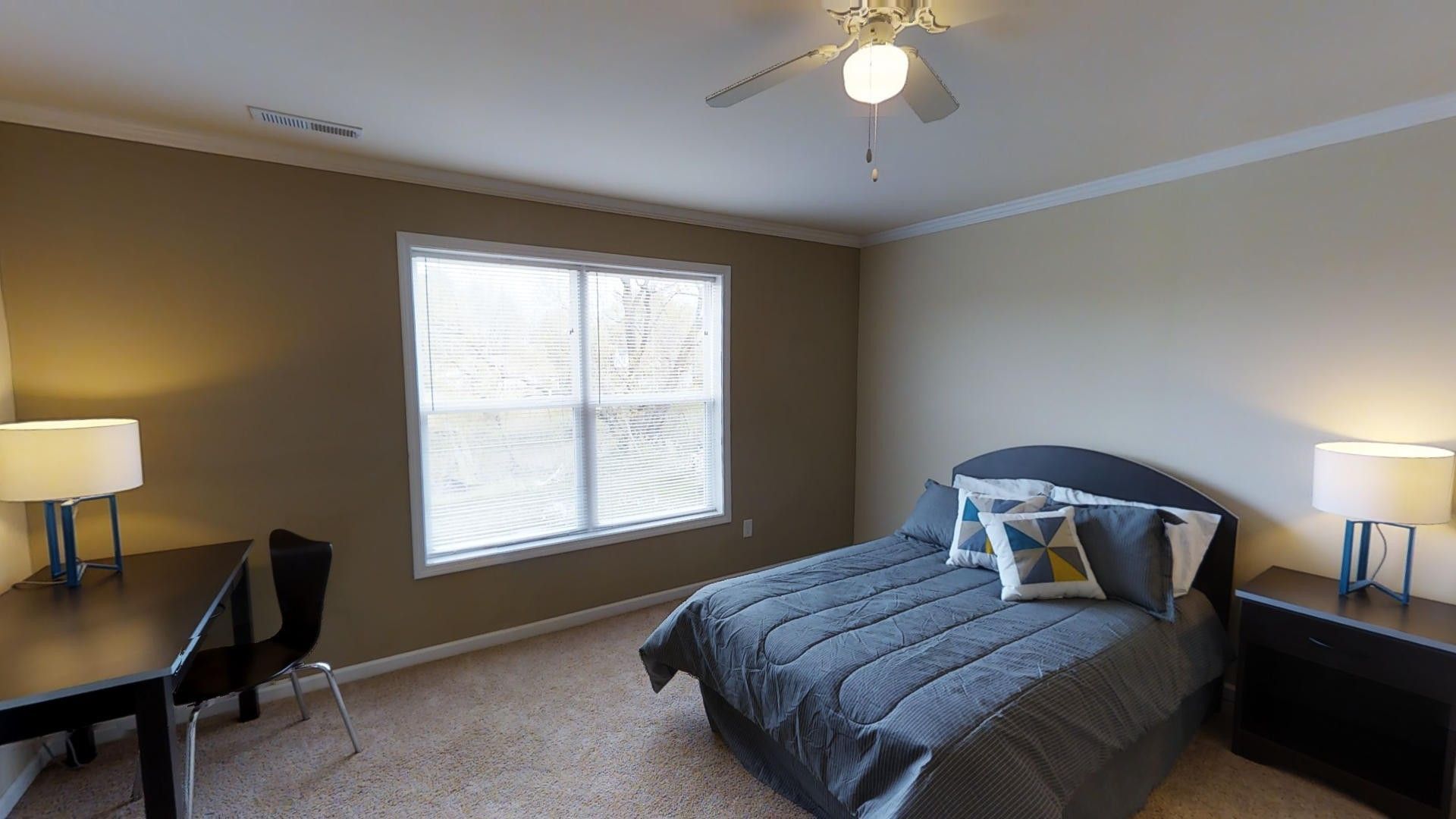 A bedroom with a bed , desk , lamps and a ceiling fan.