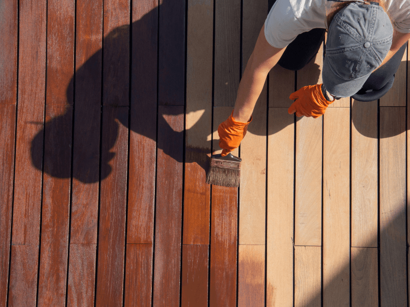 Person staining a wooden deck with a brush, wearing gloves and a hat; sunlight casts a shadow.