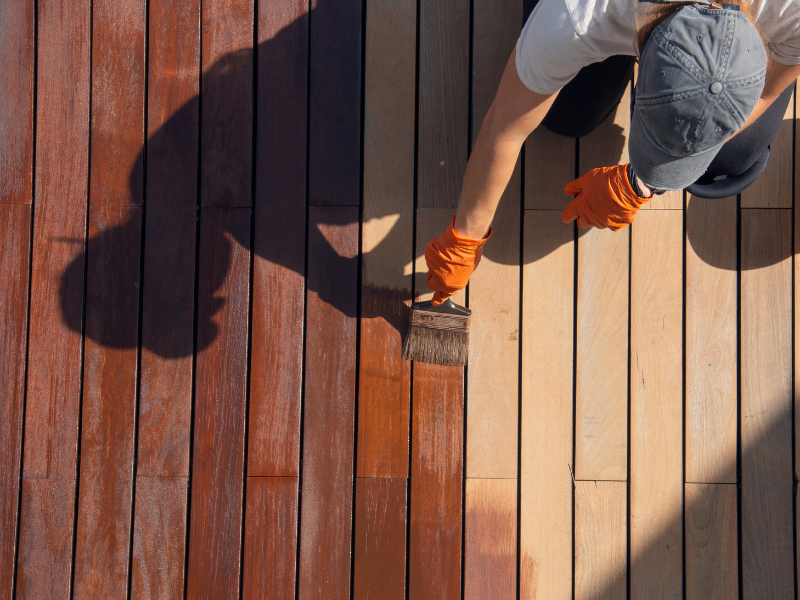 Person staining wooden deck with a brush, wearing gloves and a cap.