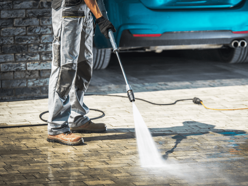 Person power washing a paved surface near a teal car at a house.