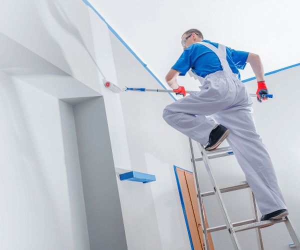 Painter on ladder using roller to paint white ceiling and wall of a house interior.