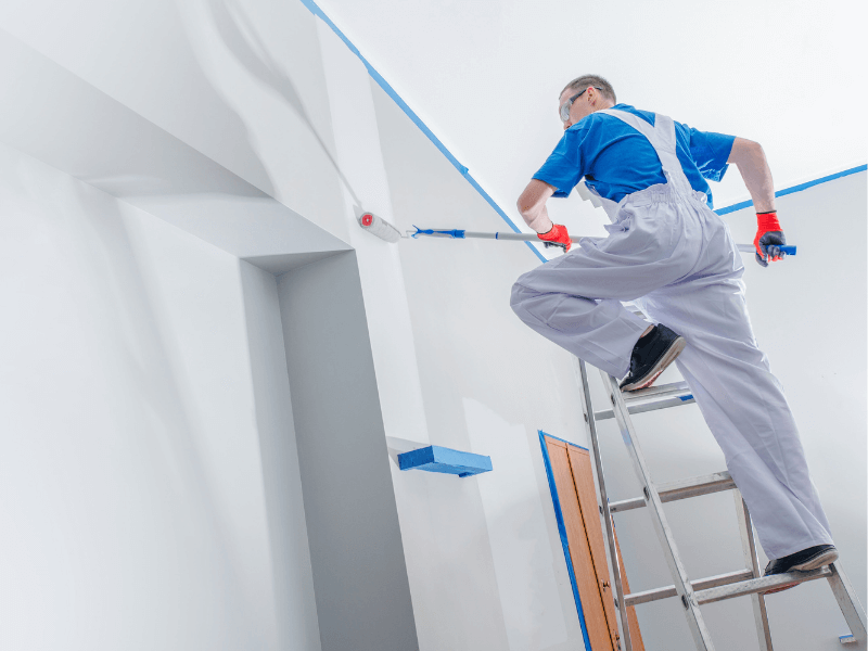 Painter on ladder using roller to paint white ceiling and wall of a house interior.