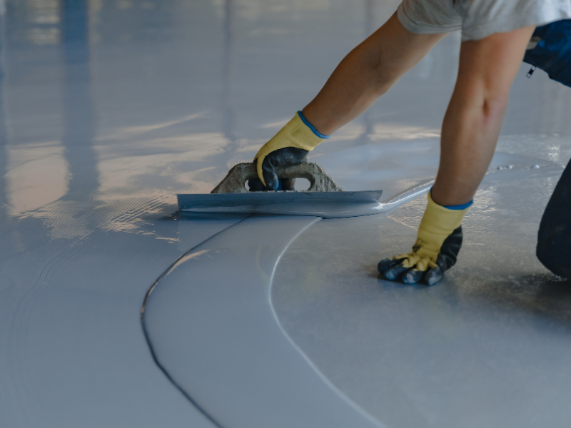 Person applying a light gray epoxy coating to a garage floor with a trowel.