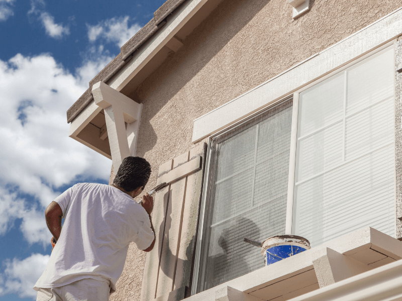 Man painting exterior trim of a house with a blue sky background.