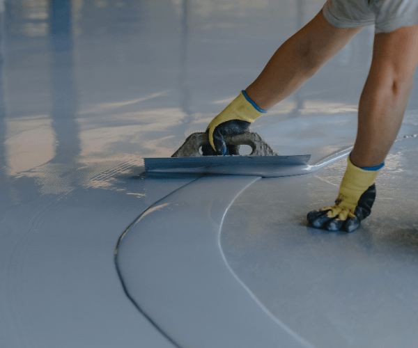 Person applying a light gray epoxy coating to a garage floor with a trowel.