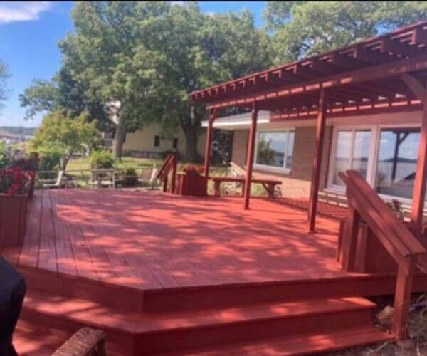 Red stained wooden deck with pergola and steps, overlooking a yard and trees.