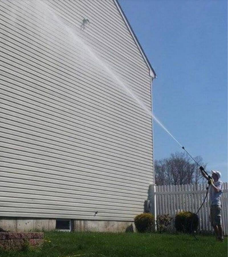 A person power washing the side of a light-colored house with white siding under a blue sky.