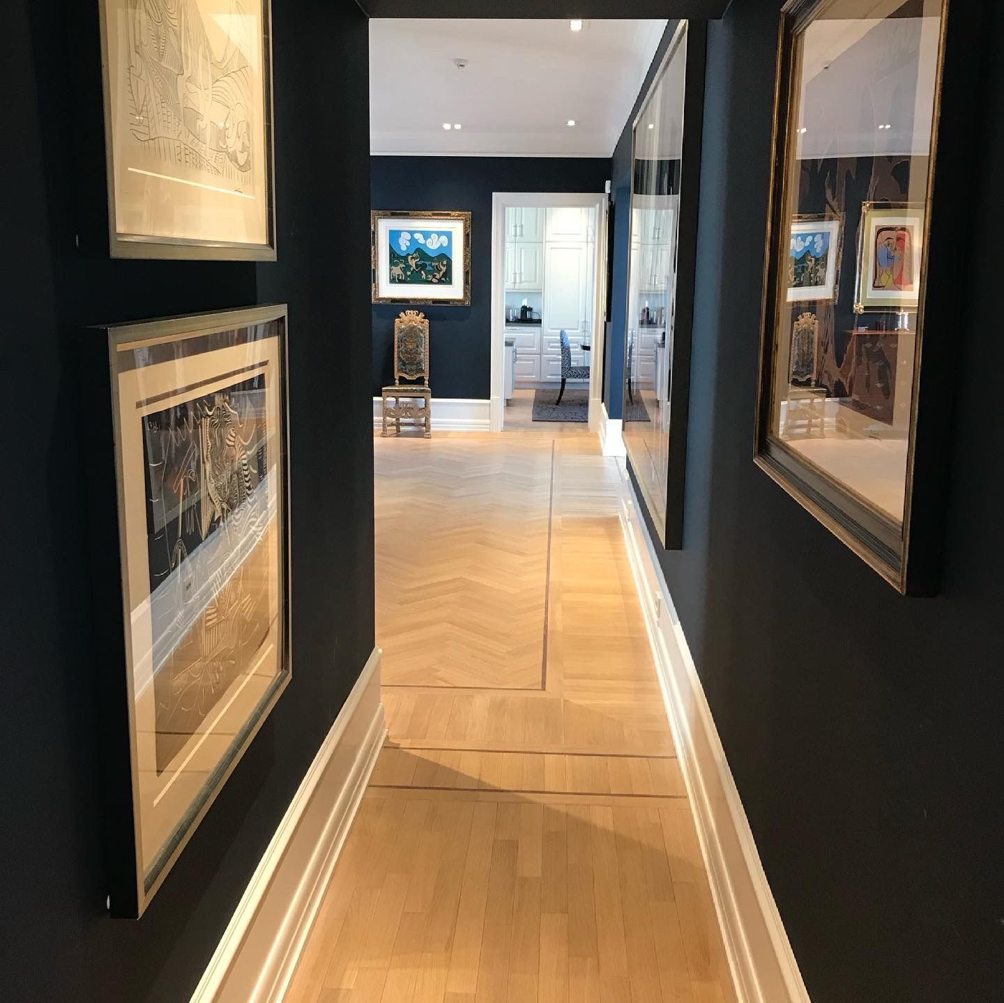 Smal gang med mørkeblå vegger, lyst tregulv og innrammet kunst. Ytterligere rom synlig i enden.