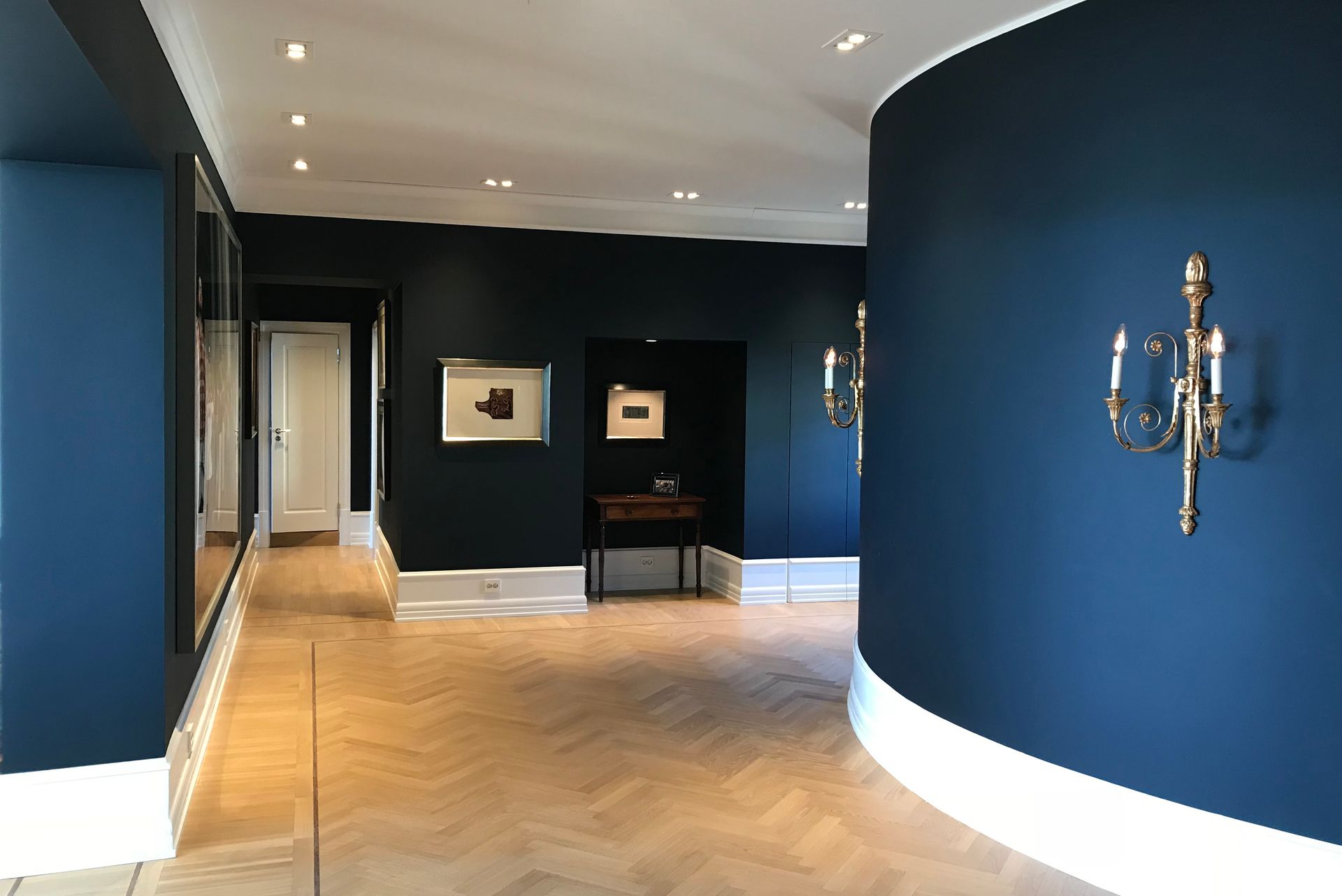 Etter-bilde. Blåvegget gang med kunstverk, fiskebensgulv og lamper.