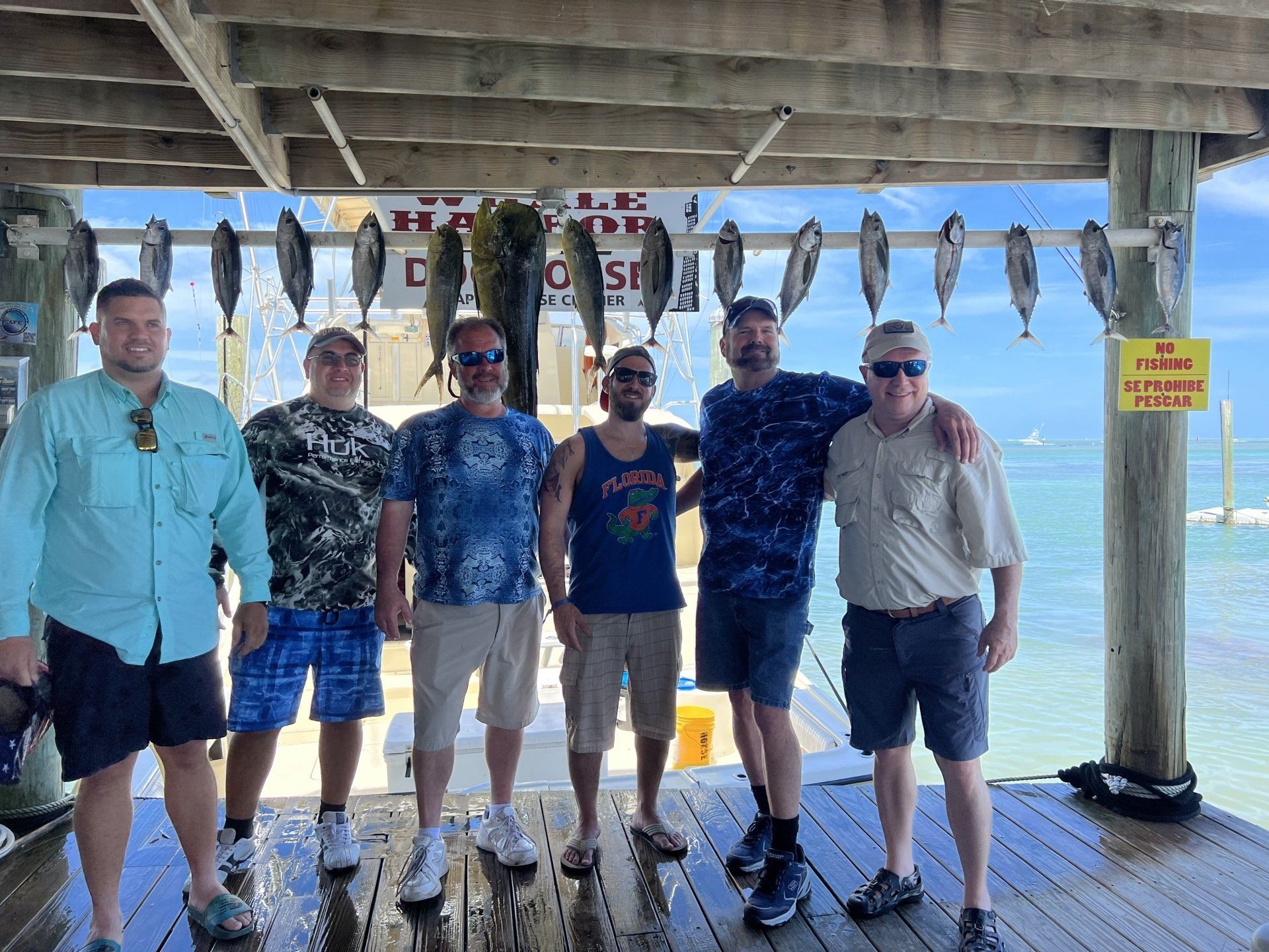 A group of men are posing for a picture on a dock with fish hanging from the ceiling.