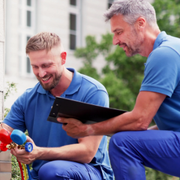 Two workers in blue uniforms inspect an outdoor air-conditioning unit with tools.